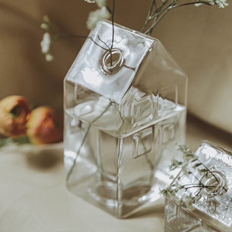 Vaso de vidro em forma de casa com flor rosa e folhas verdes