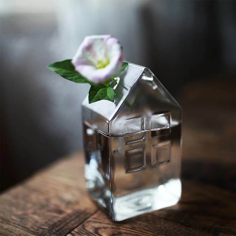 Vaso de vidro em forma de casa com flor rosa e folhas verdes