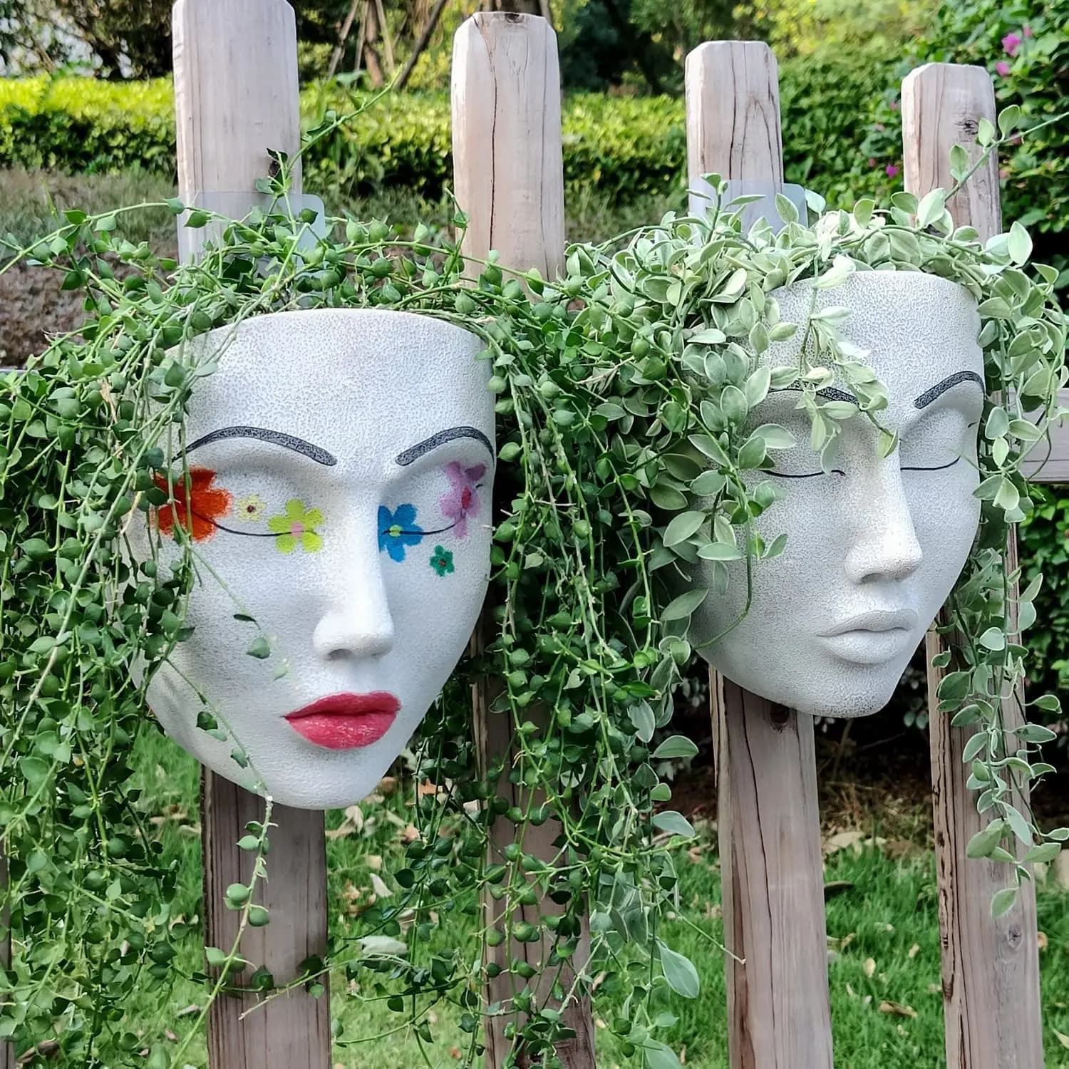 Duas máscaras de rosto humano penduradas em uma cerca de madeira com plantas verdes