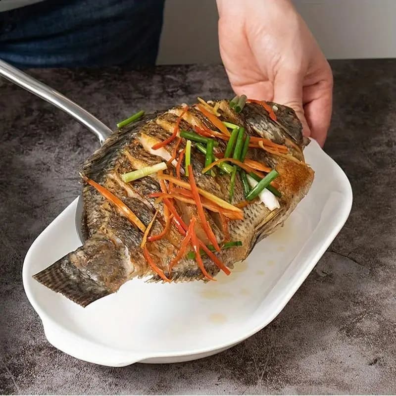 Espátula de cozinha grande em aço inoxidável segurando peixe frito decorado com legumes em prato branco