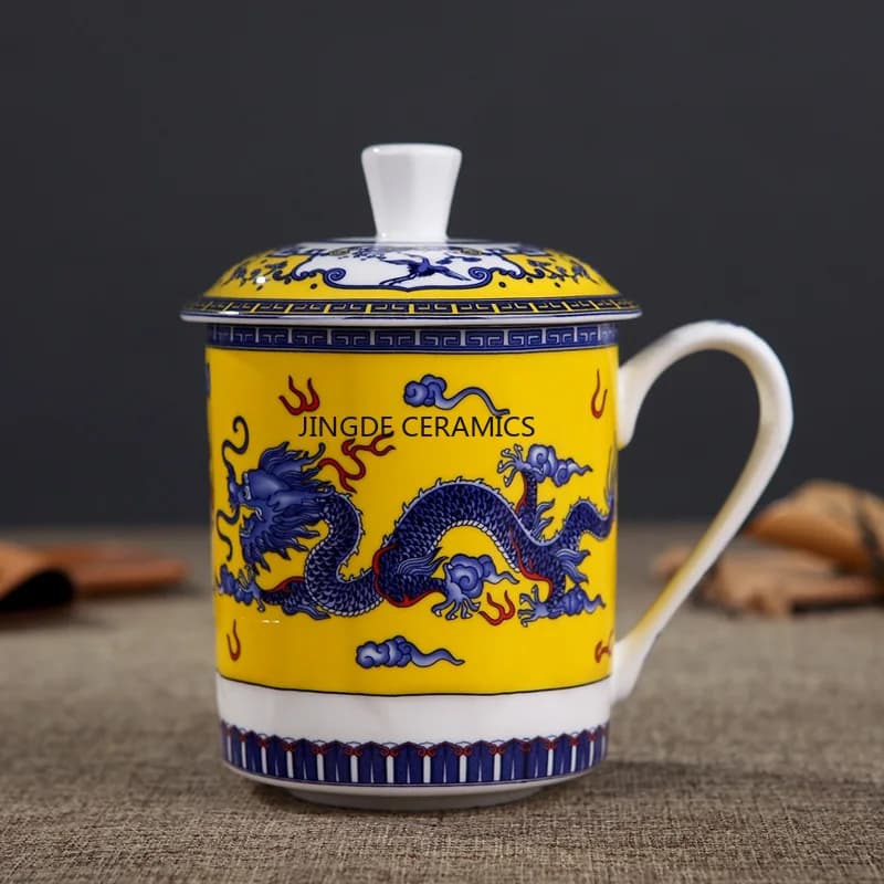 Caneca de porcelana azul e branca com tampa e alça, decorada com paisagens e personagens. Fundo cinza. A marca 'JINGDE CERAMICS' está visível na parte inferior da imagem.