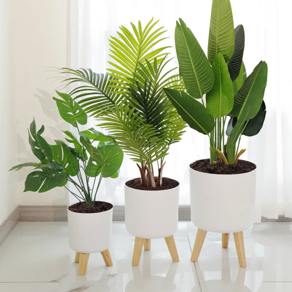 Vasos de plantas artificiais com base redonda branca e pernas de madeira em um ambiente interno.