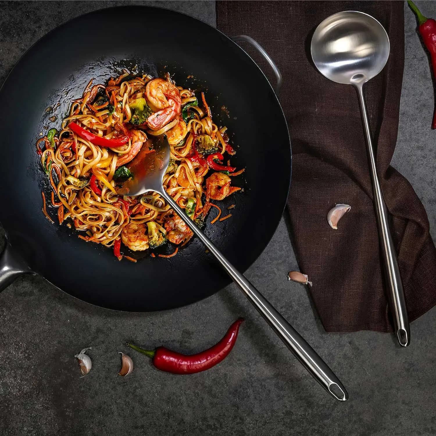 Conjunto de utensílios wok inox com espátula e concha ao lado de wok com legumes e camarão