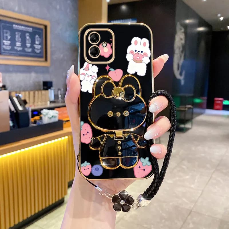 Capa de telefone preta com personagens de coelhos e frutas, estilo cartoon, detalhes dourados e alça preta, segurada por uma mão em um balcão de cafeteria.