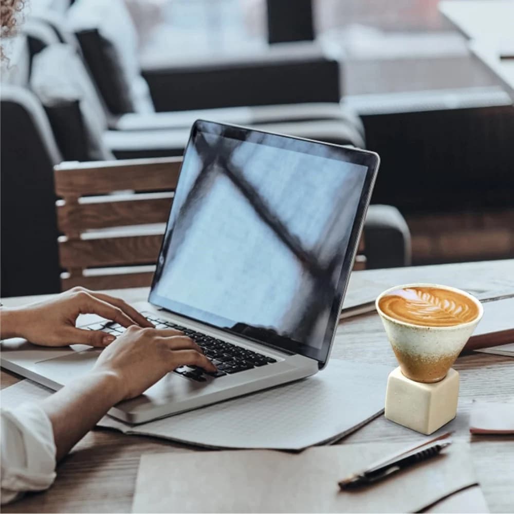 Caneca de café estilo japonês com base quadrada bege em mesa ao lado de notebook e caderno