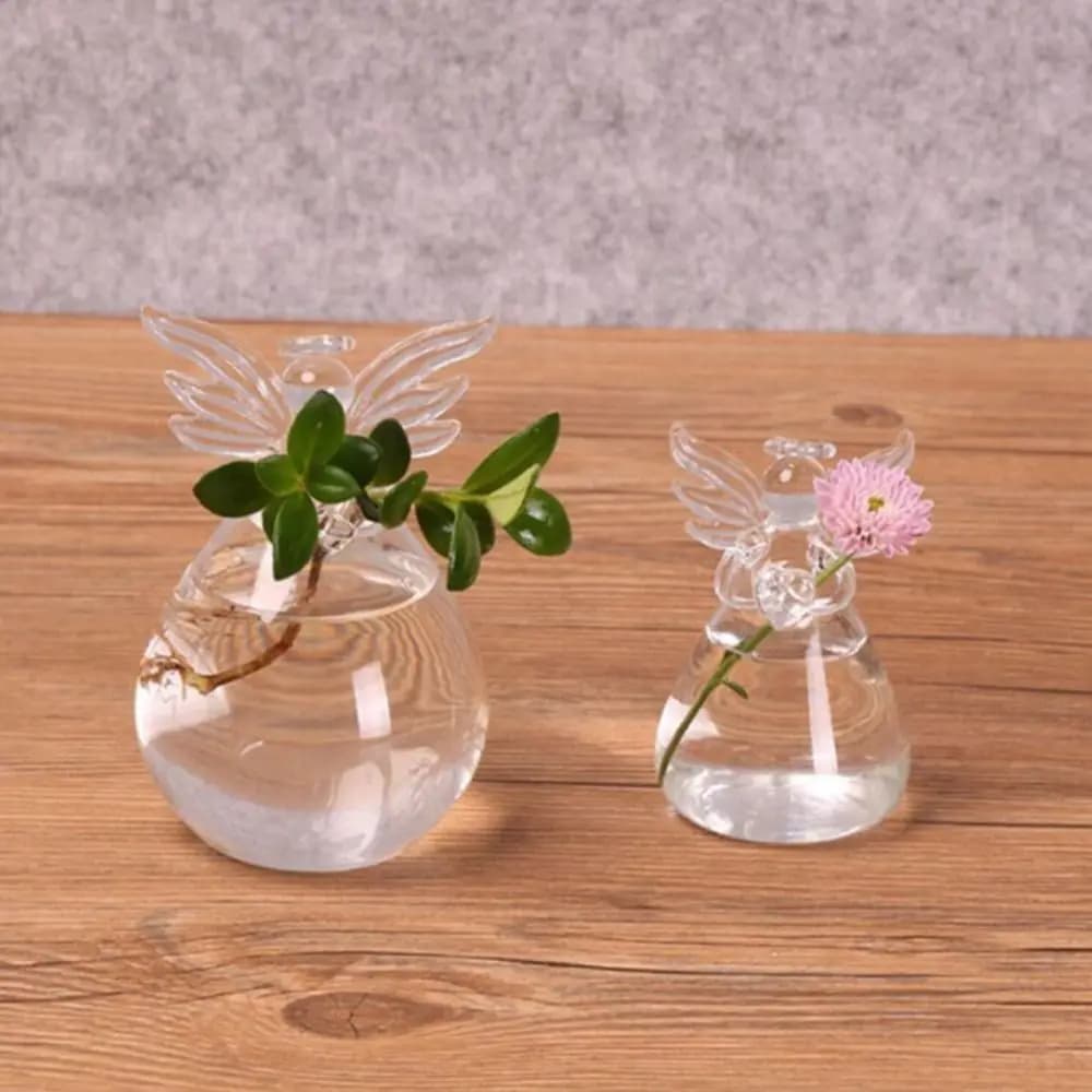 Dois vasos decorativos de vidro em formato de anjo com plantas e flores sobre mesa de madeira