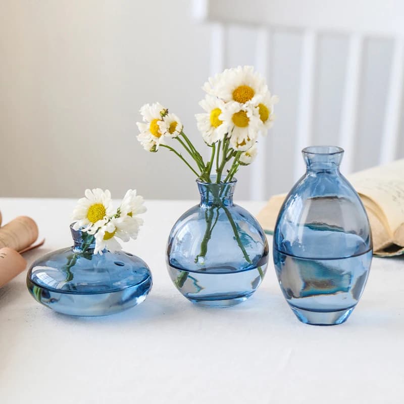 Três vasos de vidro transparente com flores brancas e amarelas em uma mesa branca