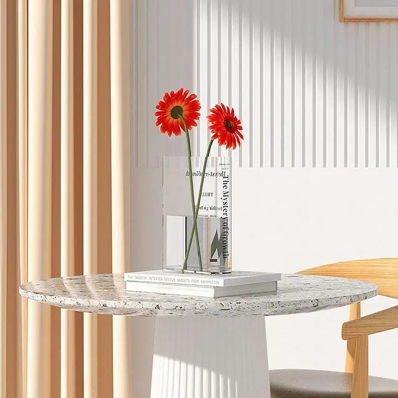 Vaso decorativo acrílico transparente formato livro com flores vermelhas sobre mesa redonda de pedra