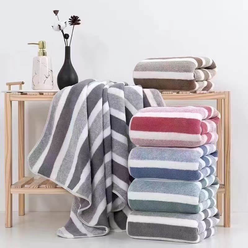 Toalha de banho coral de veludo adulto, absorvente e macia, listrada, cinza e branca, em uma prateleira.