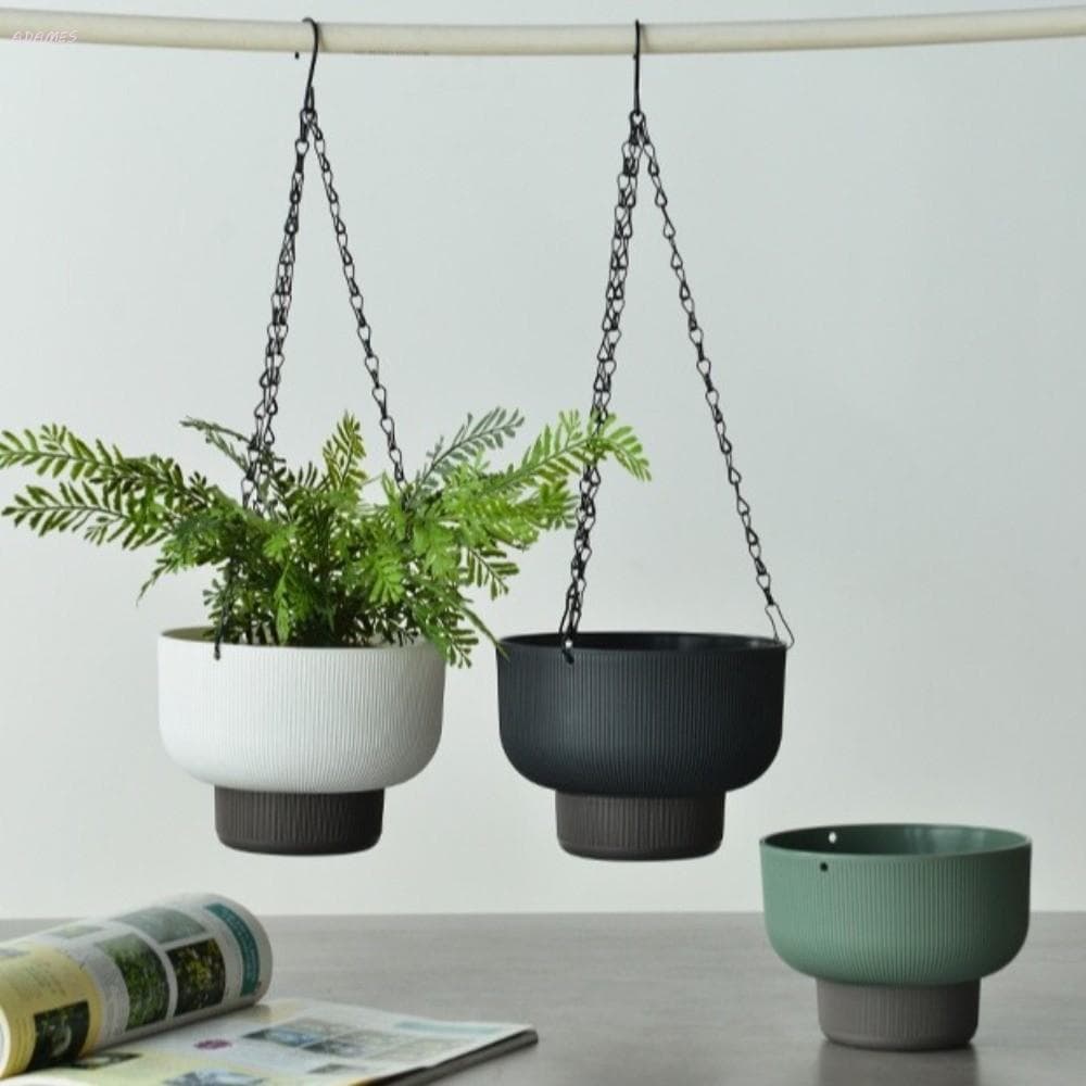 Tres vasos de flores suspensos com ganchos de corrente, dois com plantas e um vazio, ao lado de uma revista.