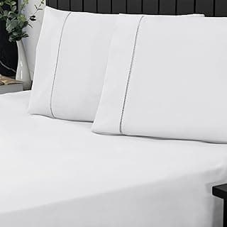 Jogo de cama king size branco em percal 400 fios, com detalhes de ponto palito, fronha e colcha branca, cama contra parede escura, planta em vaso branco.