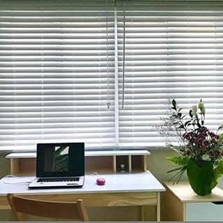 Persianas horizontais brancas fechadas com laptop aberto e flores em um vaso verde na mesa de trabalho