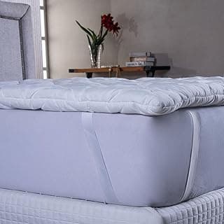 Protetor de colchão branco com toque de seda, colocado sobre colchão de cama, com fundo de parede bege e mesa com livros e flor vermelha.