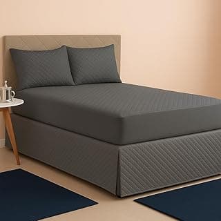 Colchão com capa impermeável cinza, cama com cabeceira estofada, dois travesseiros e tapetes azuis no quarto.