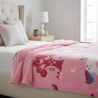 Manta infantil rosa com unicórnio e estampa de estrelas e luas, em fundo branco, sobre cama com cabeceira estofada.