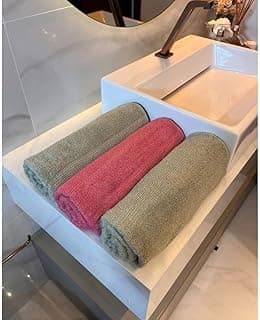 Kit de três toalhas de banho gigantes em algodão, uma rosa e duas verdes, dobradas em lavatório branco.